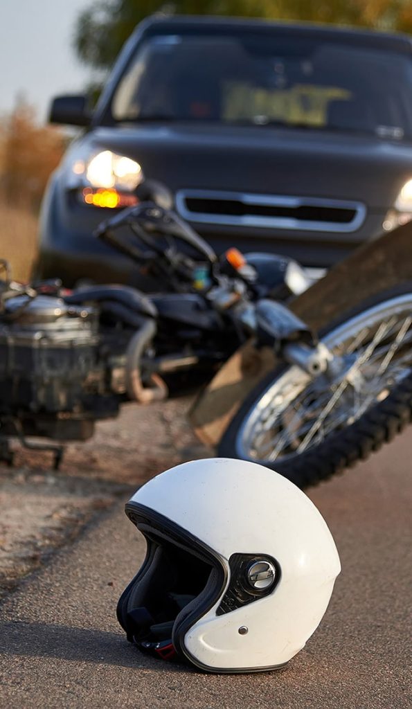 A white motorcycle helmet lies on the road, with a fallen motorcycle and a car in the background.